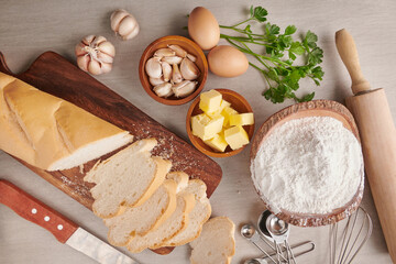 Homemade tasty bread with garlic, cheese and herbs on kitchen table. Baguette with herb butter and Rosemary, parsley and garlic with raw materials over wooden background, top view