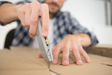 man using an utility knife to open a cardboard box - obrazy, fototapety, plakaty
