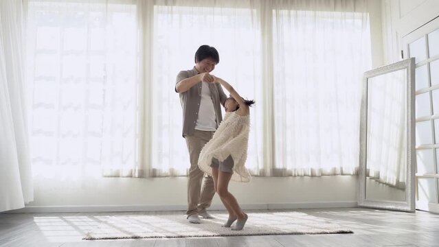 Portriat of cheerful Asian father and little daughter in beautiful dress dancing waltz, standing next to window at home. Loving korean dad and little girl having fun together at home.
