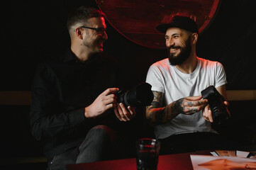 Successful professional photographers in trendy clothing sitting in cafeteria and discussing ideas for creating pictures content enjoying collaboration, happy hipster guys with slr camera communicate