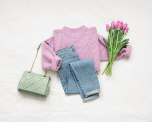 Lilac knitted sweater, blue jeans, green bag and bouquet of tulips flowers lie on white background....