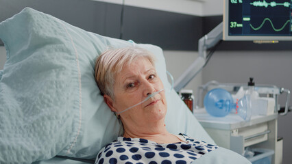 Portrait of elder patient with oxygen tube laying in hospital ward bed. Woman with illness looking...