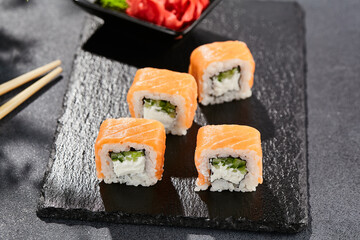 Maki sushi on dark stone table. Philadelphia maki with salmon. Sushi roll with cheese, cucumber inside, salmon outside. Style concept japanese menu with black background, leaves and hard shadow.