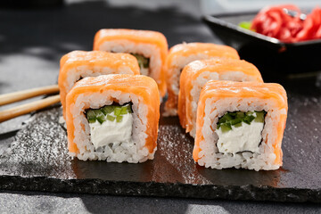 Maki sushi on dark stone table. Philadelphia maki with salmon. Sushi roll with cheese, cucumber inside, salmon outside. Style concept japanese menu with black background, leaves and hard shadow.