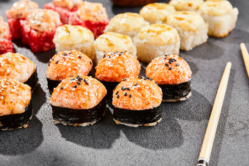 Hot maki set on dark slate. Baked sushi set with variety rolls. Sushi rolls with salmon,tobiko, crab and baked cheese topped. Style concept sushi menu with black background, leaves and hard shadow.