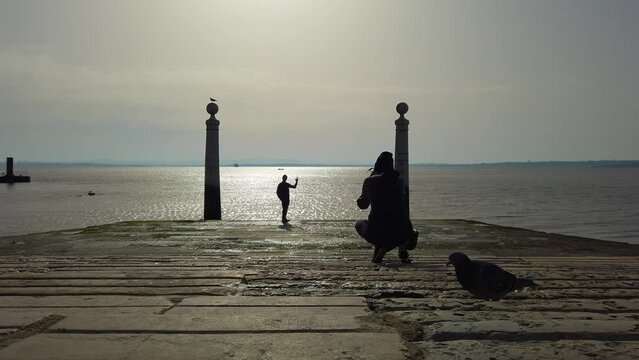 Person taking photo of posing friend, Tagus river in background, silhouette back view, Cais das Colunas, Lisbon, Portugal