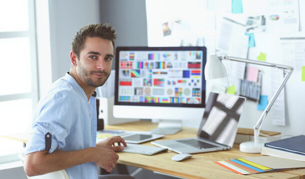 Portrait of young designer sitting at graphic studio in front of laptop and computer while working online.