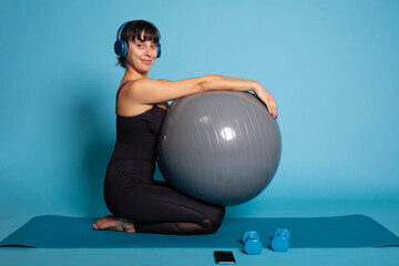 Personal trainer listening music using headphones while working at wellbeing stretching body muscles in studio. Athlete woman in sportwear leaning on yoga fitball. Sport concept