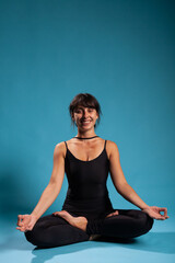 Obraz premium Athletic woman meditating during gymnastics workout sitting in padmasana position on yoga mat practicing spiritual meditation. Gymnast adult training body muscles in studio with blue background