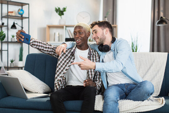 Positive same sex couple in casual wear taking selfie on modern smartphone while resting together on comfy couch. Happy loving people spending time with fun at home.