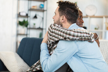 Caucasian and african american men sitting together on couch in strong hugs. Connection, relations...