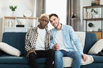 Obraz na płótnie Canvas Happy and beautiful gay couple smiling and looking at camera while sitting in embrace together on couch. Two multiracial men in casual clothes posing at bright living room.