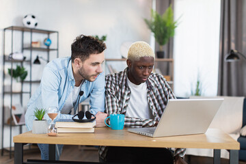 Obraz premium Multiracial same sex couple of two handsome gays using modern laptop ai living room. African american man sitting at desk while caucasian standing near.