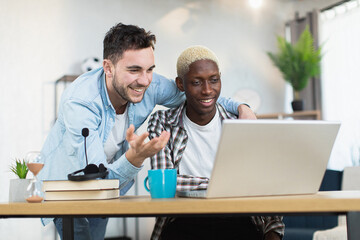 Smiling caucasian man embracing his african american boyfriend that sitting at desk and using wireless laptop. Two loving gays surfing internet during free time at modern apartment.
