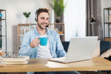 Obraz premium Smiling young man in casual wear drinking tea during video chat at home office. Caucasian male person using modern laptop and headset for remote work.