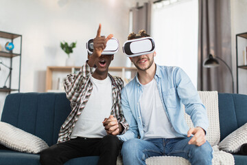 Happy same sex couple of two multiracial men using VR goggles during leisure time at home. Handsome young gays sitting on couch and holding hands.