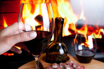 Female hand hold wine glass against wine table with appetizers for wine and electric fireplace. Manicure with red hearts. Romantic dinner at home concept.