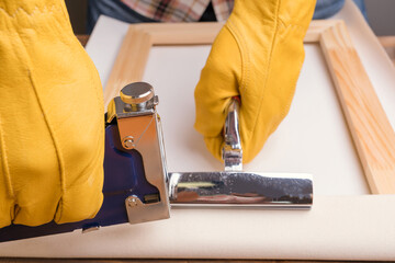 The artisan's hands in yellow protective gloves pull the canvas onto a wooden stretcher using a...