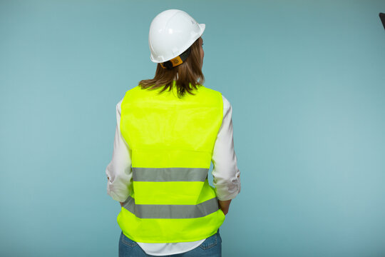 Professional engineer. A woman in a protective helmet and a bright vest stands with her back to the camera, against a blue background. Copy paste.