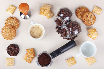 Cup of espresso, crackers, cookies, holder with ground coffee, tamper and cans of coffee beans on table. View from above.