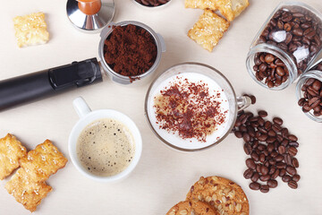 Cup of espresso, cappuccino with chocolate crumbs, crackers, cookies, holder with ground coffee, tamper and coffee beans on table. View from above.