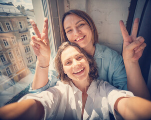 Young beautiful lesbian couple making selfie on smartphone while sitting on windowsill in street view apartment