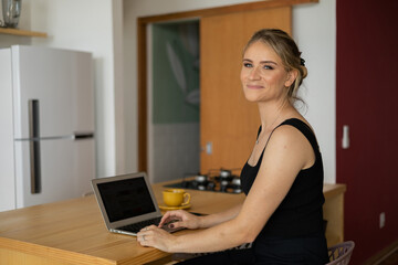 Young woman working at home in her kitchen with laptop and papers on kitchen wooden desk. Home office concept.