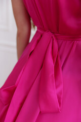 Portrait of a young brunette Caucasian woman with long straight hair in a bright pink dress. Revealed thigh-high slit. In the background is a white wall and a fireplace