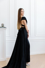 Fototapeta premium stylish and gorgeous girl in a black glittery dress poses in a photo studio against a white wall. The slit of the dress at the hip and the beautiful body shape.