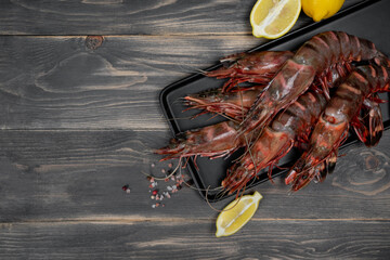 Raw black tiger shrimps on black plate and wood table, decorated with lemon slices.