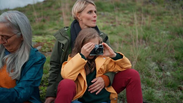 Small girl with mother and grandmother taking pictures with camera on top of mountain.