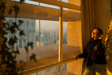 Fototapeta premium Side view of young woman listening to music with headphones in her ears by the window with a city view landscape in the background. High quality photo