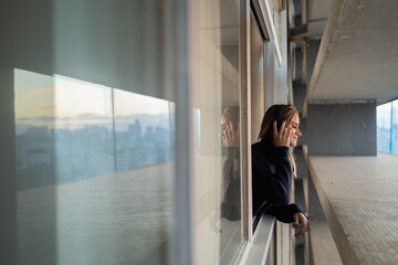 Side view of young woman listening to music with headphones in her ears by the window with a city view landscape in the background. High quality photo