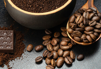 ground coffee in a cup with a spoonful of coffee beans and a bar of dark chocolate on a black background