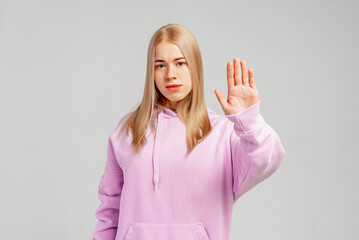 Serious blonde girl showing stop sign and frowning displeased, telling no, refusing something, standing in trendy hoody over gray background