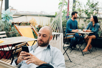 shaved head boy in the hammock spends time with his mobile phone on an outdoor terrace - hostel bar...