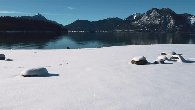 Snow covered beach at Lake Walchensee in Bavaria, south Germany in the scenic alps mountains close to Austria. Blue clear drinking water waves at the white shore and blue sky in winter. Drone aerial