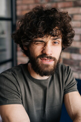 Close-up face of friendly handsome young man winking looking at camera sitting on chair in cozy living room with brick wall. Closeup vertical shot of confident curly male posing at home.