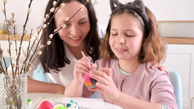 Little girl and mom paints eggs for Easter. Child of European appearance , decor on the table. Happy family makes decorations for Easter and colored eggs for the holiday. 4k 