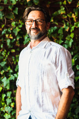 Portrait of adult mature handsome bearded man with white shirt smiling at the camera and wear eyewear. Foliage leaf green background Vertical composition. Happy male people style