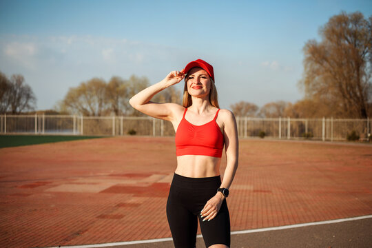 Portrait of smiling sportswoman in red cap, top and black cycling shorts outdoor on the sport ground