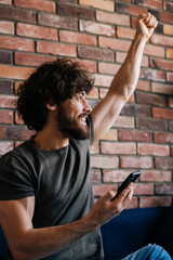 Vertical shot of cheerful excited young man looking away, receive online message with good news on...
