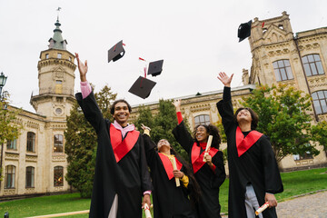 Multiracial graduates wearing gown laughing while tossing their caps
