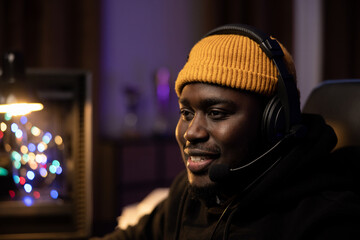 The gentle smile of a man spending the night in front of his computer. A guy with a yellow cap on his head and a headset is playing games. Dark room lit by colored led lights.