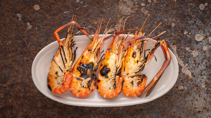 grilled giant freshwater prawns in oval ceramic plate on rusty texture background, top view, flat...