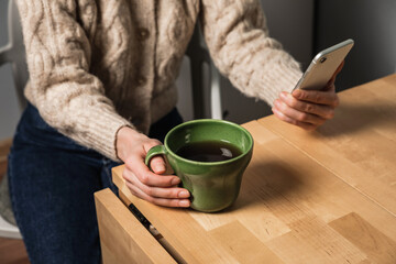 A caucasian woman in a cozy beige cardigan using smartphone, sitting at the table and drinking tea