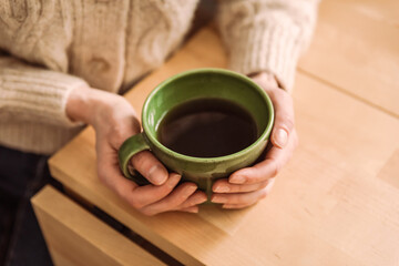 Fototapeta na wymiar A caucasian woman in a cozy beige cardigan holding a green cup of tea in her hands, sitting at the table, a close-up natural light lifestyle image