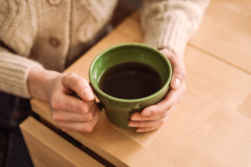 A caucasian woman in a cozy beige cardigan holding a green cup of tea in her hands, sitting at the table, a close-up natural light lifestyle image