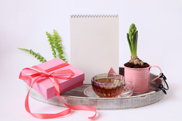 Morning hot tea on a holiday: blooming hyacinth, a cup of tea, a notepad for a congratulatory inscription, a gift tied with a pink ribbon on a decorative tray, white background