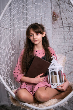 A cute little girl is reading a book sitting in a hanging chair. Portrait of a child studying at home.
